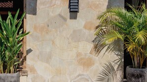 En stenmur med ett litet ventilationsgaller, omgiven av två krukväxter och en träaltan.