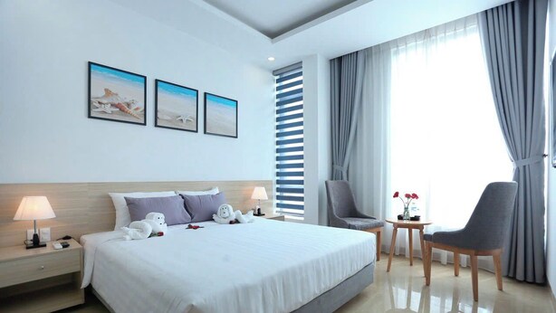 A modern hotel room with a large bed, two bedside tables, a chair, a small table with flowers, and a large window with curtains. - Basic Hotel Ha Long (Bai Chay)