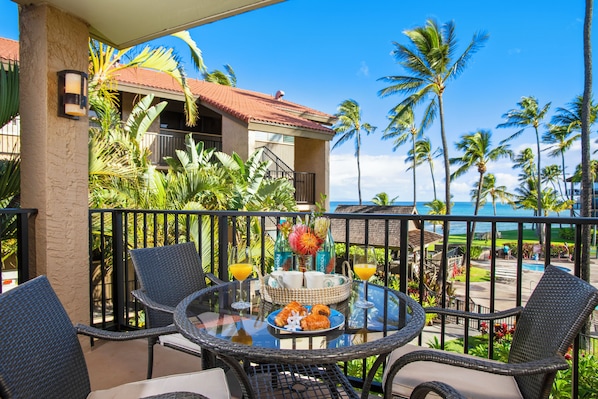 A balcony with wicker chairs, a glass table, and a tropical view including a pool and palm trees.