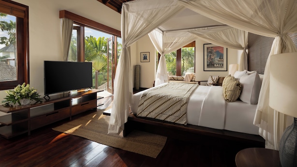 A bedroom with a canopy bed, a flat-screen TV, and a view of palm trees. - The Suar Seminyak by Nakula (Seminyak)
