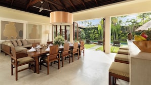 A spacious dining area with a wooden table set for six, a sofa with cushions, and a large window offering a view of a lush garden and pool. - The Suar Seminyak by Nakula (Seminyak)