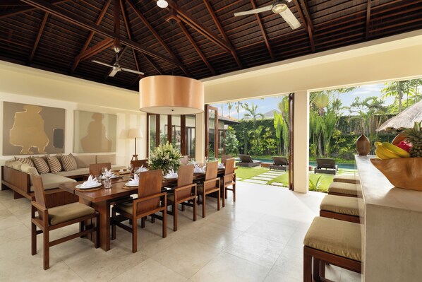 Uma ampla sala de jantar com uma mesa de madeira posta para seis pessoas, um sofá com almofadas e uma grande janela com vista para um jardim exuberante e uma piscina.