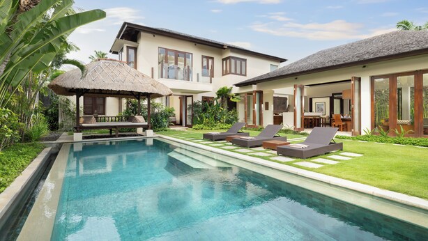 A swimming pool with lounge chairs, a thatched-roof structure, and a large house with multiple windows and doors. - The Suar Seminyak by Nakula (Seminyak)