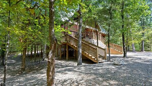 Une cabane en bois avec un escalier menant à une terrasse, entourée d’arbres et bordée d’un sol en gravier.