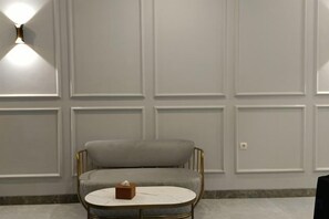 A room with a grey upholstered chair, a round table with a tissue box, and a wall with paneling. - D'Ninety Six Inn (Playen)