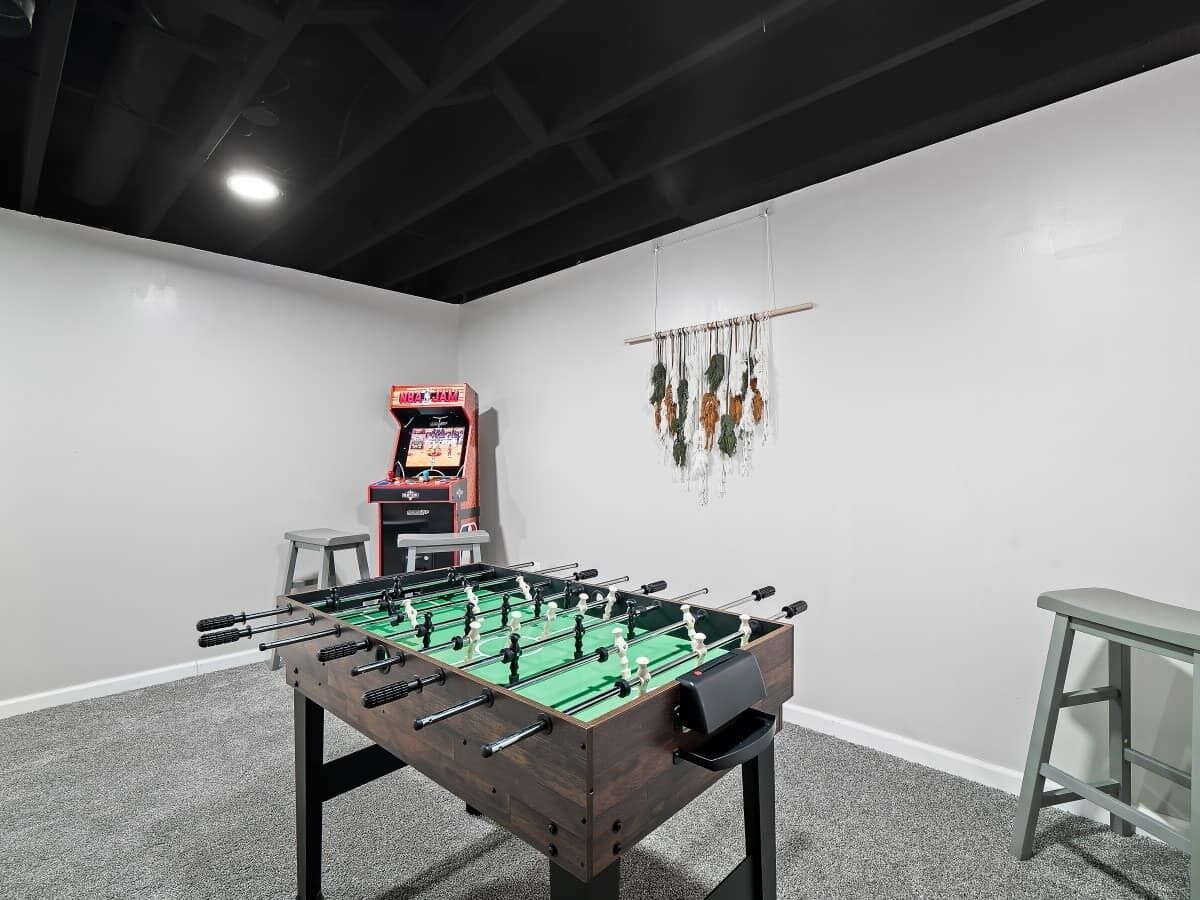 A room with a foosball table, a small stool, and a foosball machine against the wall.