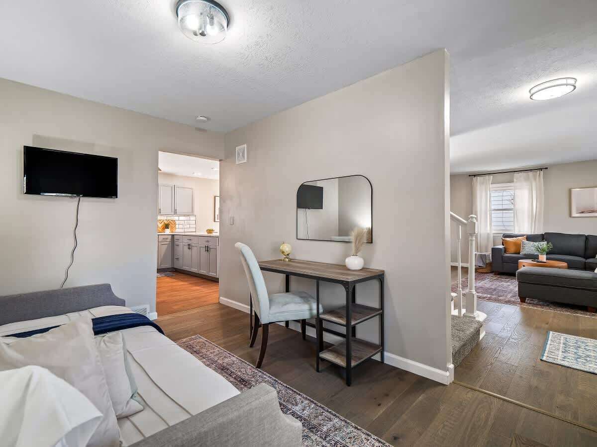 A modern living room with a sofa, a dining table with a chair, a TV mounted on the wall, and a staircase leading to another room.