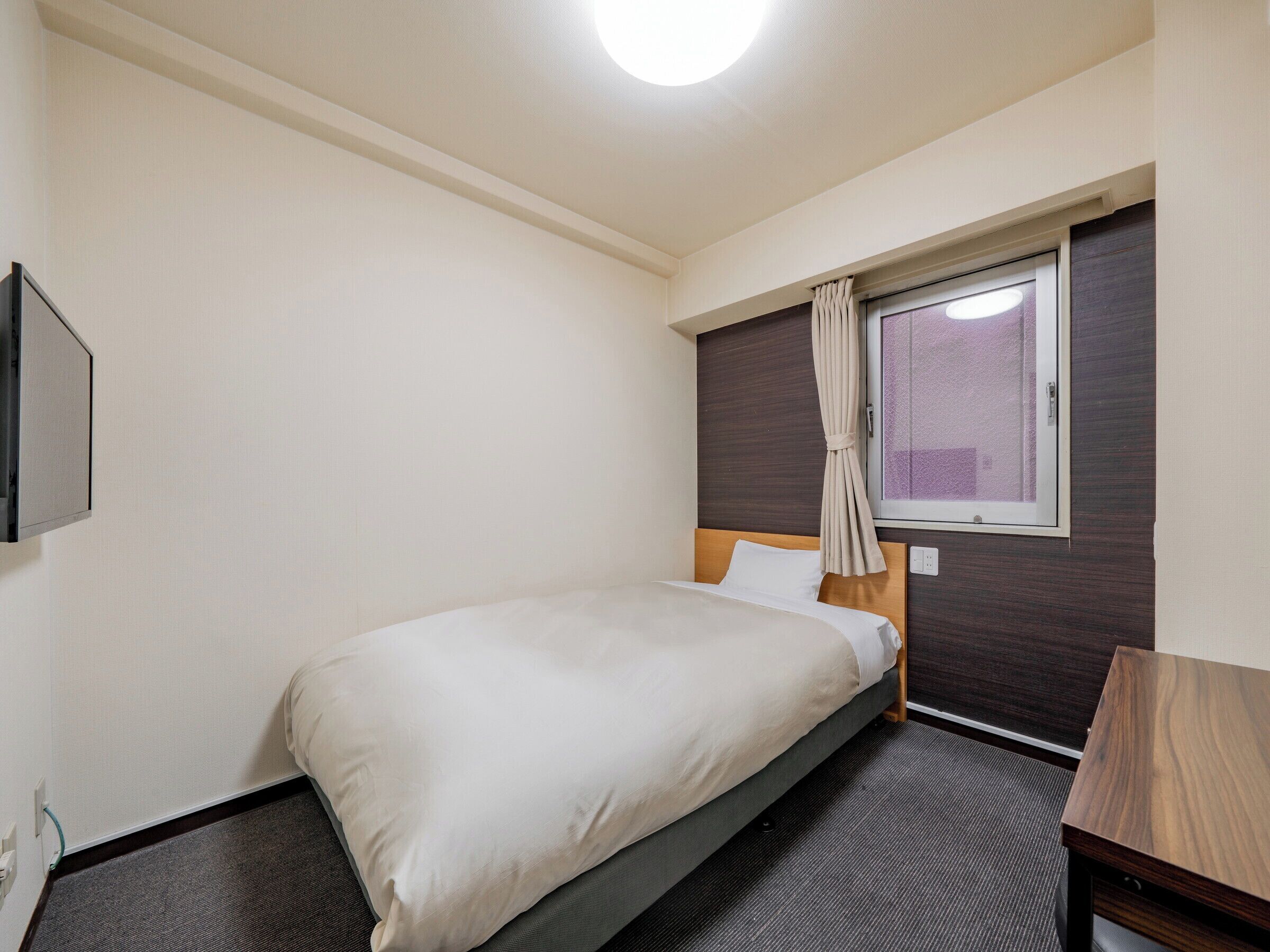 Ein kleines, ordentlich eingerichtetes Hotelzimmer mit einem Einzelbett, einem Nachttisch aus Holz, einem Fenster mit Vorhängen und einem an der Wand befestigten Fernseher.