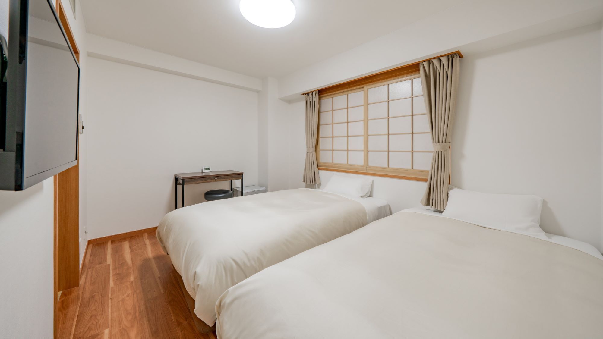 A hotel room with two beds, a small table with a clock, a TV mounted on the wall, and a window with curtains.
