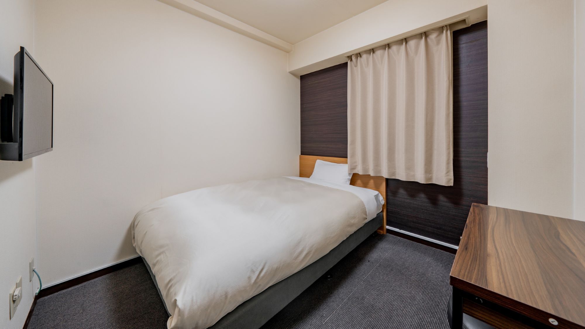 A hotel room with a single bed, a wooden headboard, a dark carpet, a small wooden desk, and a wall-mounted TV.