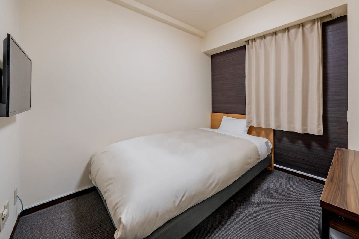 Habitación de hotel con una cama individual, cabecera de madera, alfombra oscura, escritorio de madera pequeño y televisor montado en la pared.