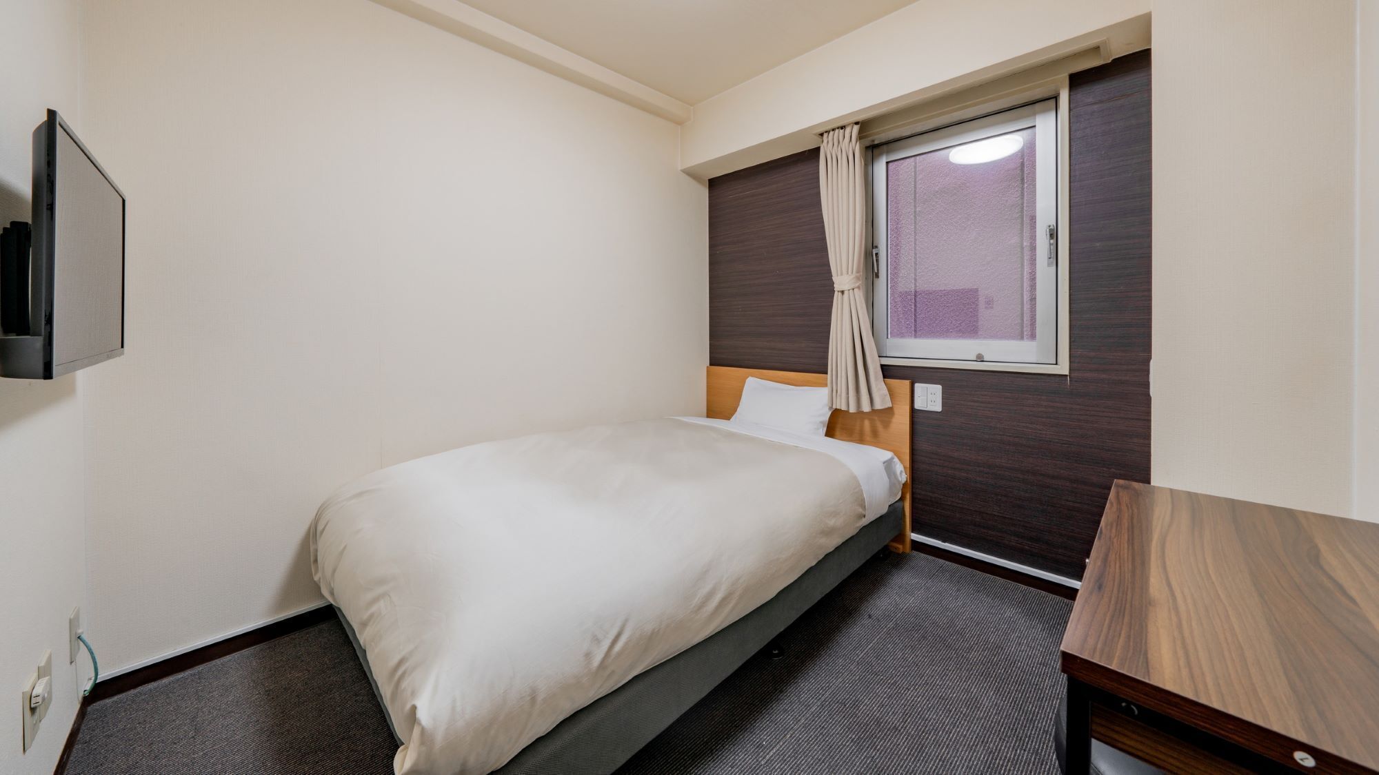 A single bed with a wooden headboard, a small bedside table, a window with curtains, and a wall-mounted TV.
