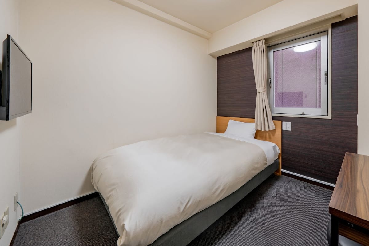 Una cama individual con cabecera de madera, una mesita de noche pequeña, una ventana con cortinas y un televisor montado en la pared.