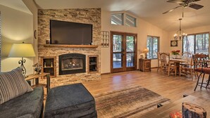 A spacious living room with a stone fireplace, a flat-screen TV mounted above it, and a dining area with wooden furniture. - Bobcat Bungalow - Quaint, Quiet & Cozy Retreat in Munds Park - Yes, there's A/C! (Munds Park)
