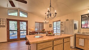 A spacious kitchen with a large island, wooden cabinets, and a chandelier. There are two windows with blinds, a door leading to a deck, and a refrigerator. - Bobcat Bungalow - Quaint, Quiet & Cozy Retreat in Munds Park - Yes, there's A/C! (Munds Park)