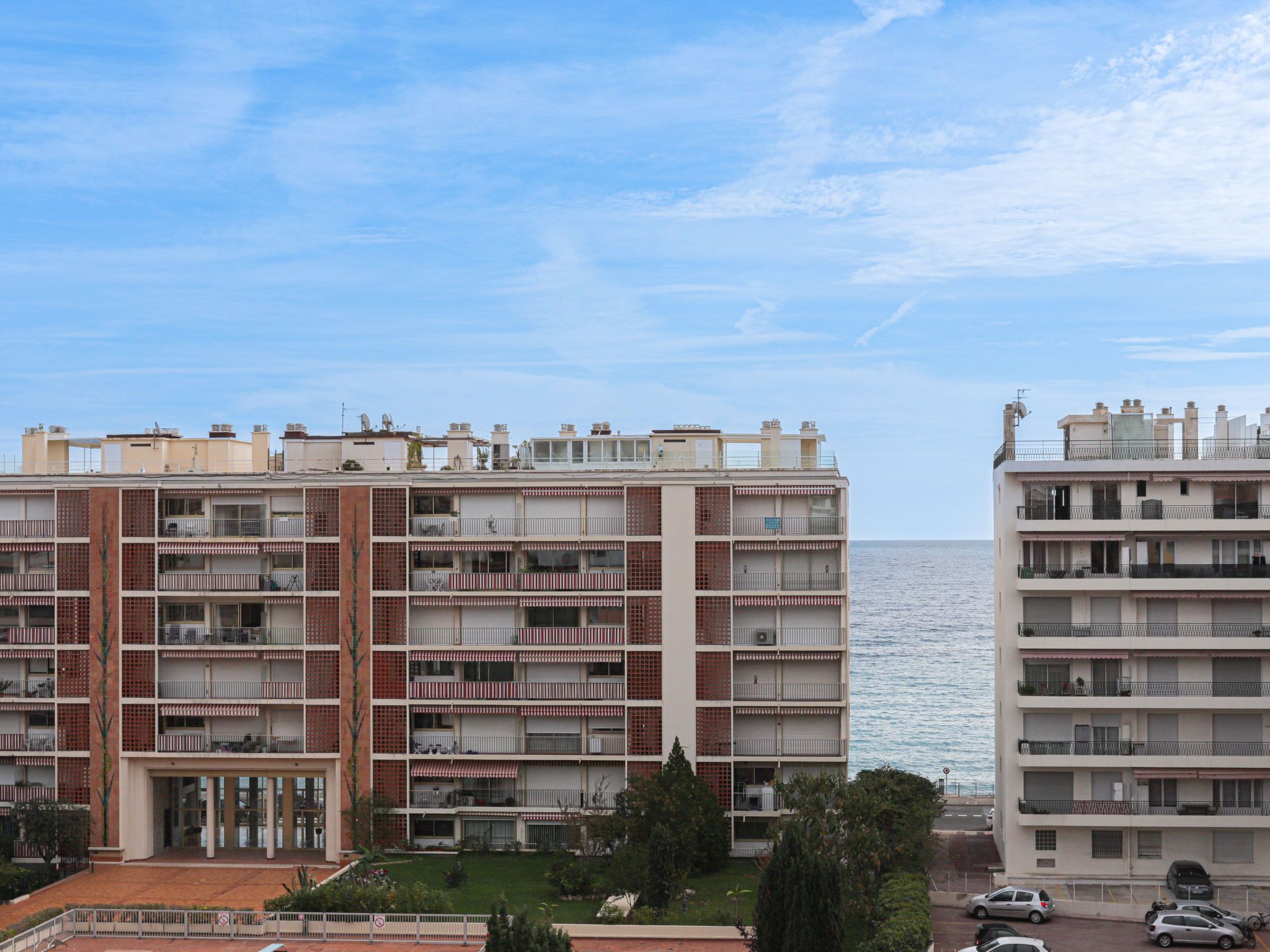 A multi-story residential building with balconies, a parking area, and a view of the sea.