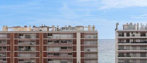 A multi-story residential building with balconies, a parking area, and a view of the sea.