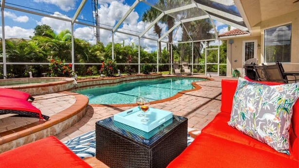 Une piscine extérieure couverte, dotée de chaises longues rouges et blanches, d’une table basse en verre et offrant une vue sur une végétation tropicale.