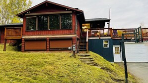 Una cabaña de madera con terraza, garaje y un pequeño espacio exterior con un aro de baloncesto.