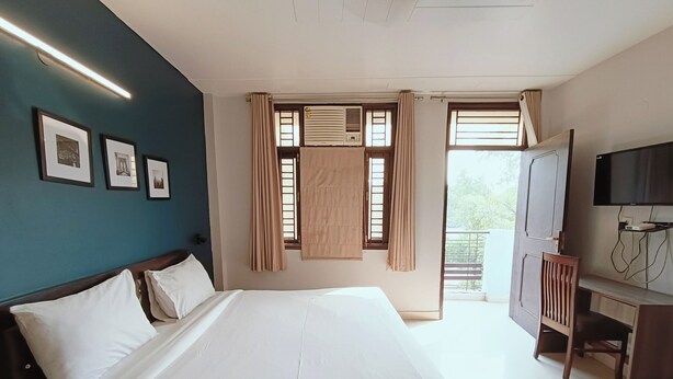 A hotel room with a large bed, a desk with a TV, and a balcony with curtains. - Silver Arrow Guest House  (New Delhi)