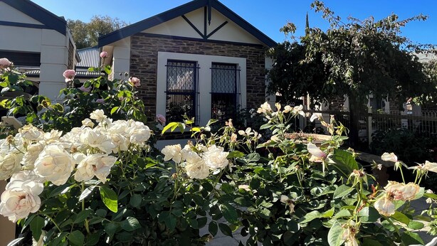 Une maison avec une façade en pierre, un toit à pignon et un petit jardin où poussent des roses blanches.