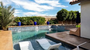 A swimming pool with two lounge chairs, a waterfall feature, and a view of a desert landscape.