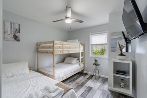 Une chambre avec un lit superposé, un ventilateur de plafond, une fenêtre avec des stores, un téléviseur fixé au mur et une petite étagère avec une plante.