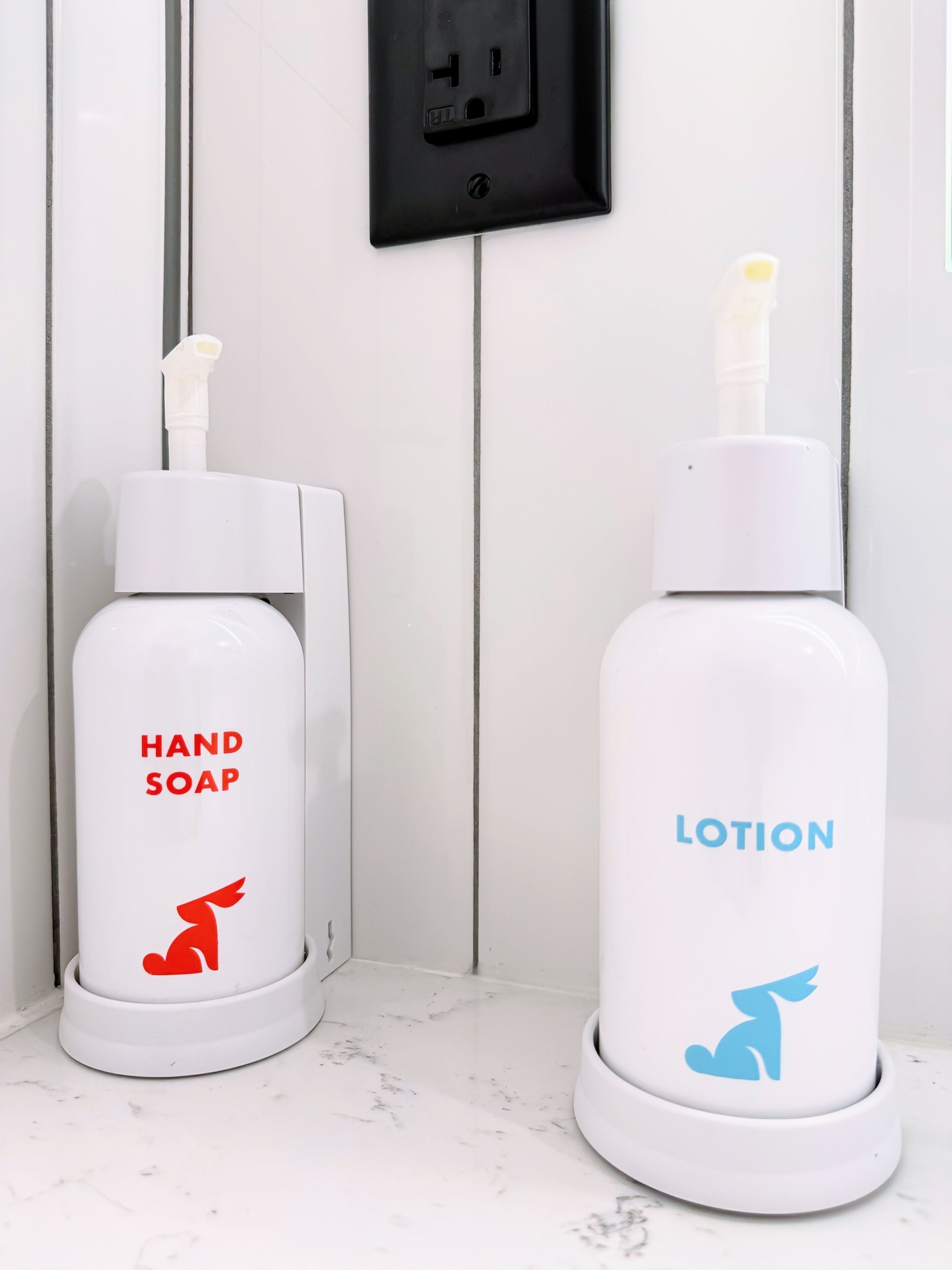 Due erogatori bianchi su un piano di marmo, uno con l'etichetta "HAND SOAP" e l'altro con "LOTION", con una presa di corrente sullo sfondo.