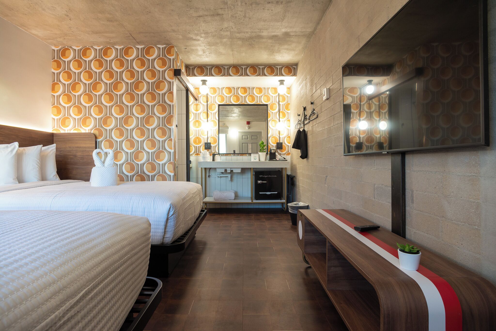 Habitación de hotel con cama, banco, televisor, espejo y baño con lavamanos y toallero.