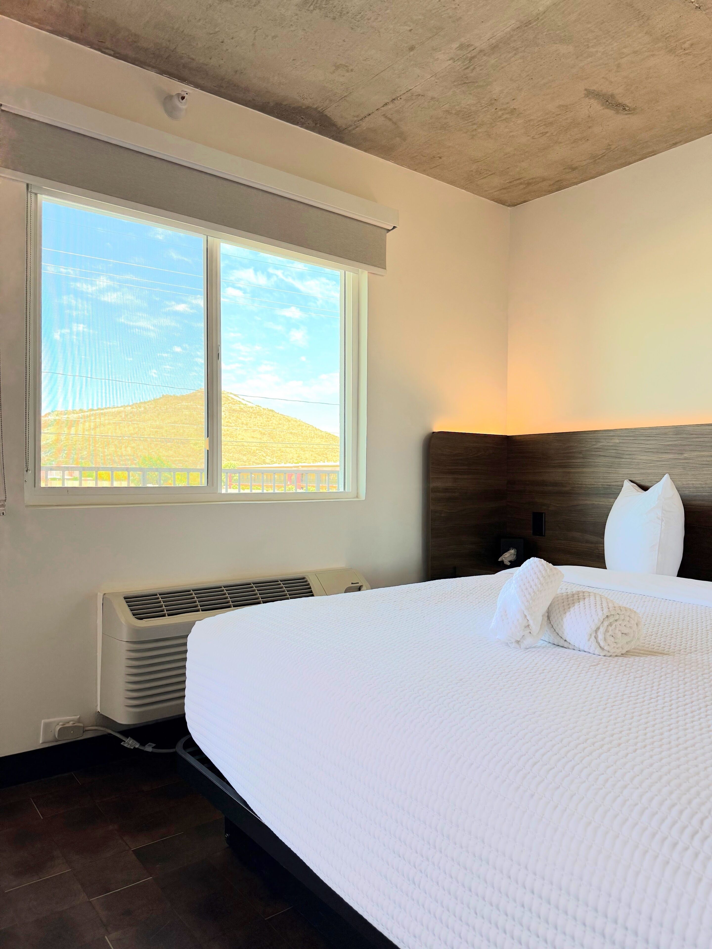 Habitació d'hotel amb un llit gran, una finestra amb vista a un paisatge amb turons, aire condicionat fixat a la paret i un panell de fusta al capçal.