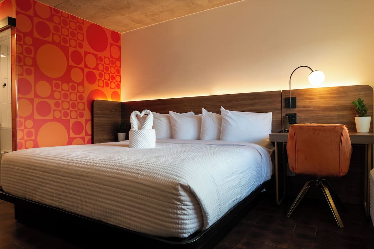 Habitación de hotel con una cama grande, cabecera de madera, mesita de noche con lámpara y una planta en maceta.