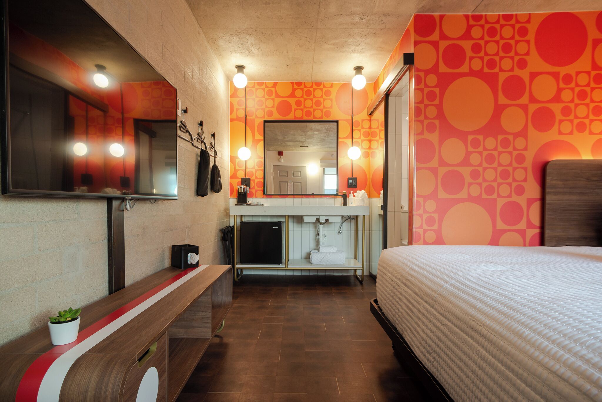 A modern hotel room with a wooden bench, a small plant, a mirror, a sink, and a bed.