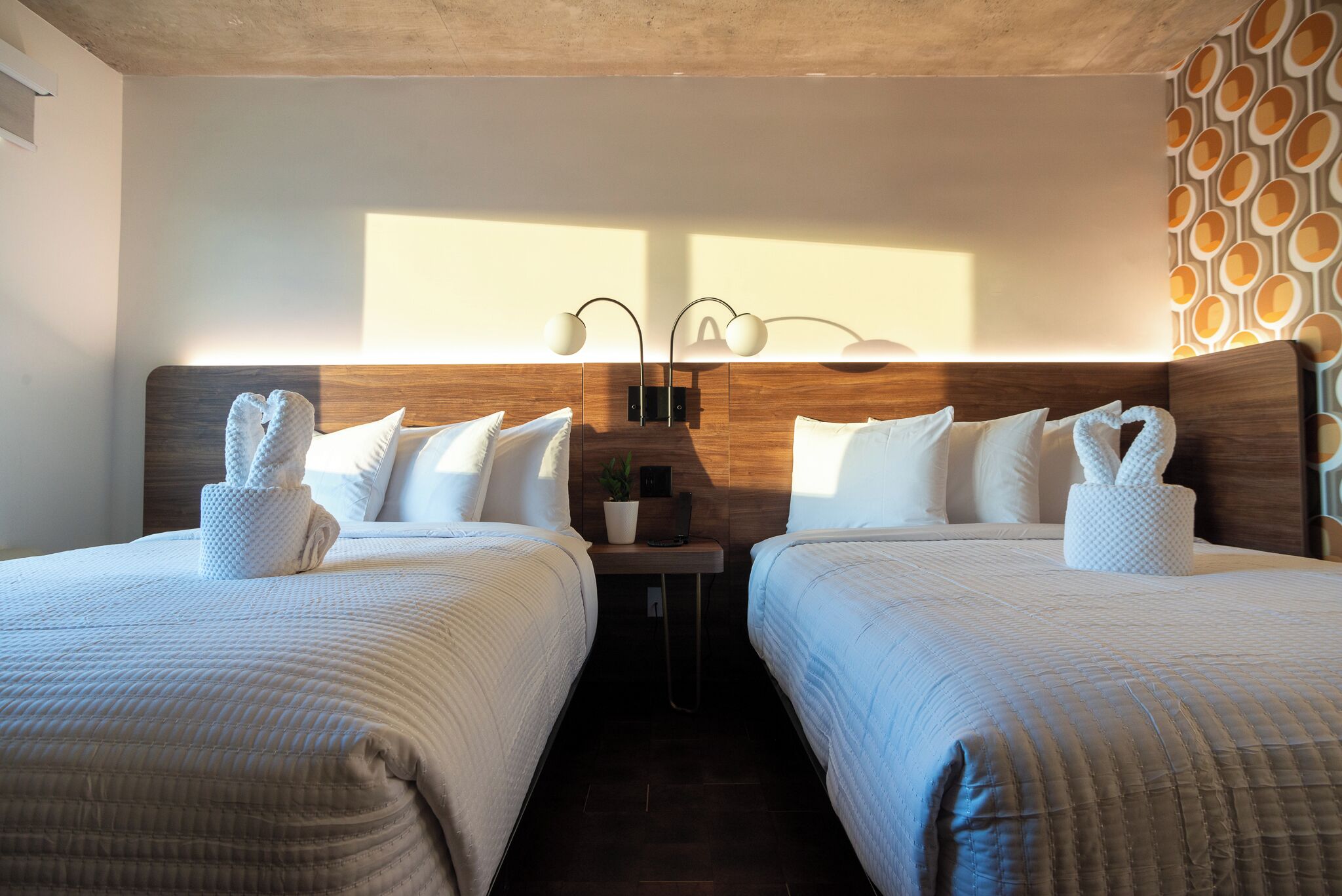 A hotel room with two beds, each with a heart-shaped towel, a bedside table with a lamp, and a wall-mounted light fixture.
