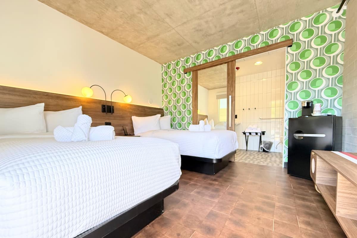 Habitación de hotel con dos camas, cabecero de madera, pared con estampado verde, refrigerador negro y una banca de madera.