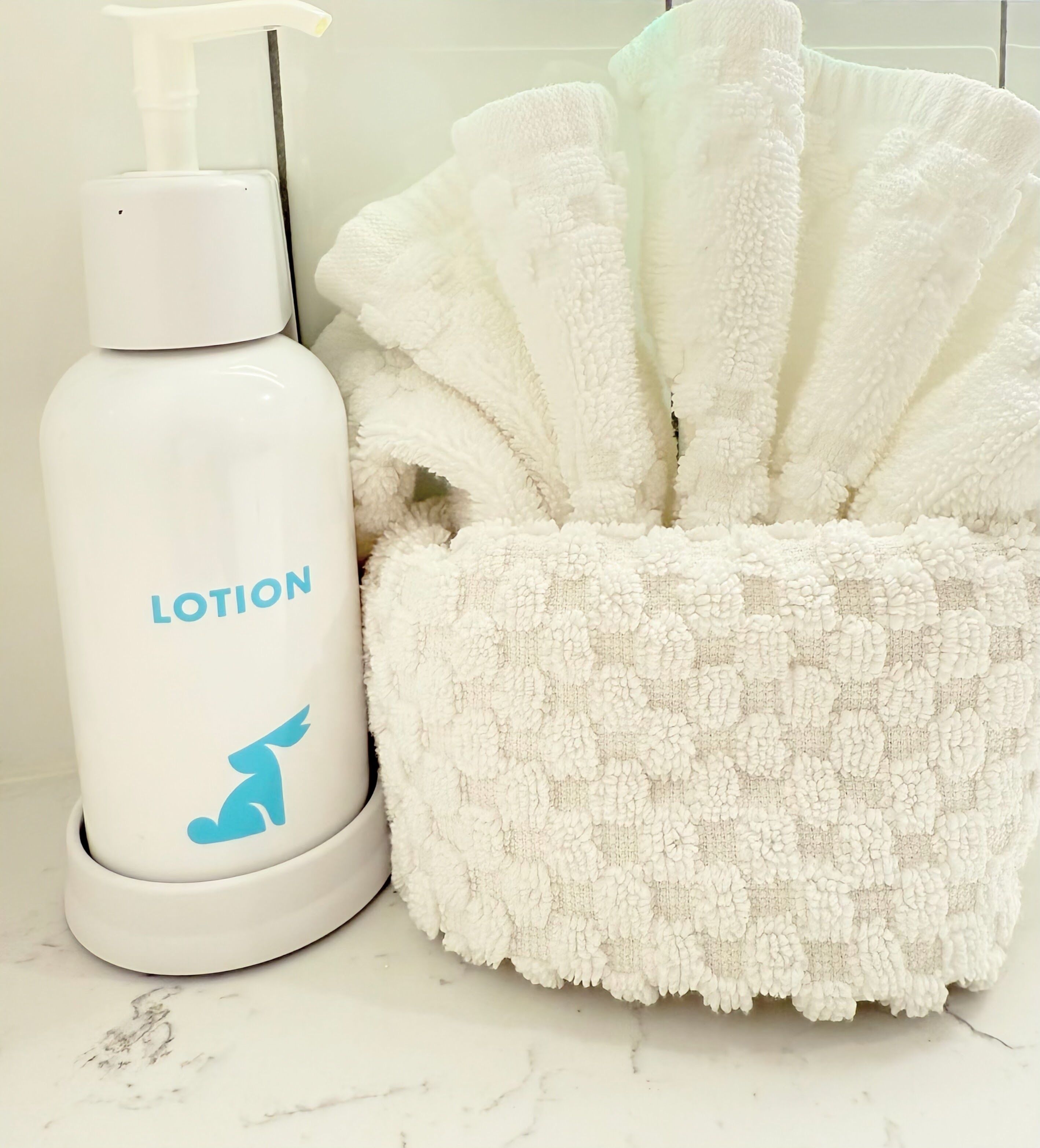 Eine weiße Lotionflasche mit Pumpspender und ein flauschiges weißes Handtuch auf einer Marmorplatte.