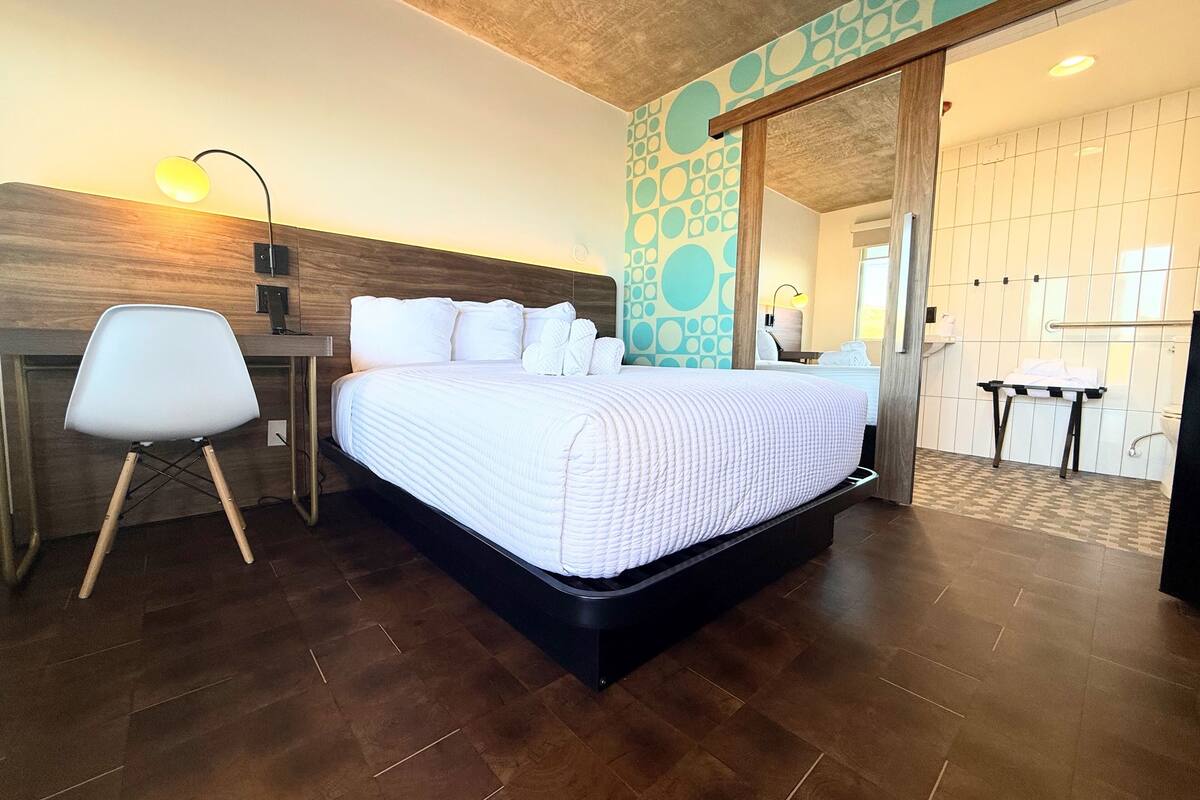 Habitación de hotel ordenada con cama, escritorio y silla. Se puede ver un baño a través de una puerta abierta.