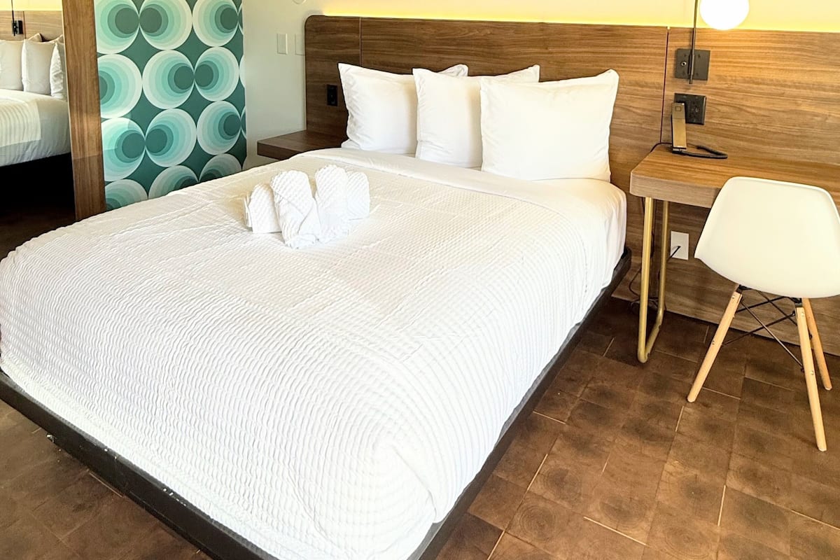 Habitación de hotel con una cama grande, cabecero de madera, mesita de noche con lámpara, una silla y un espejo.