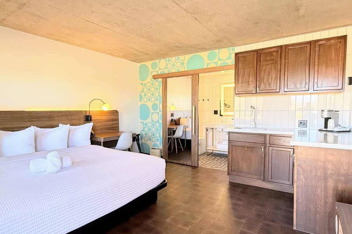 Habitación de hotel con una cama grande, un escritorio, una silla, una kitchenette y un baño visible a través de una puerta abierta.