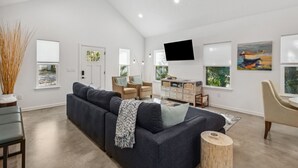 Un vaste salon doté d’un canapé d’angle gris foncé, d’une table basse en bois et d’un téléviseur à écran plat fixé au mur. On y trouve deux fenêtres avec des stores blancs, une porte et une petite table d’appoint sur laquelle est posé un vase contenant des plantes séchées.