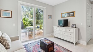 A living room with a white sofa, a coffee table, a TV on a white cabinet, and a sliding glass door leading to an outdoor patio. - American Beach Retreat - Hosted by Moore AI Rentals (Fernandina Beach)