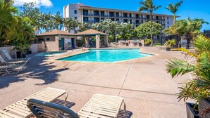 Zona de piscina hotelera con sillones, una zona de estar cubierta y un edificio con balcones al fondo.