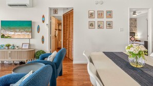 Una sala de estar moderna con un cuadro grande, una zona de comedor con mesa y sillas, y un pasillo que lleva a un dormitorio.