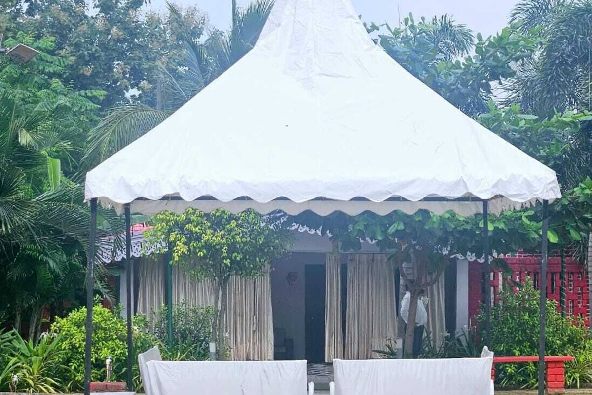 A white tent with a peaked roof, surrounded by greenery and a red building in the background.