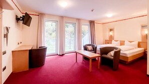 Habitación de hotel con una cama, dos sillas, un escritorio, un cesto de basura y un televisor montado en la pared.