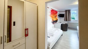 Ett hotellrum med en säng, en tv, ett fönster med gardiner, ett element och en papperskorg.