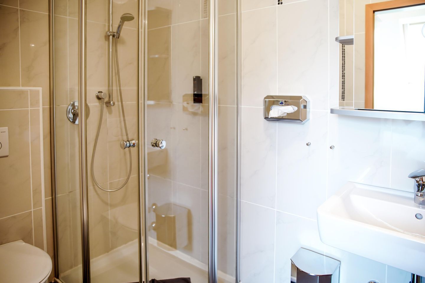 A glass-enclosed shower with a handheld showerhead, a wall-mounted towel dispenser, and a white sink with a mirror above it.
