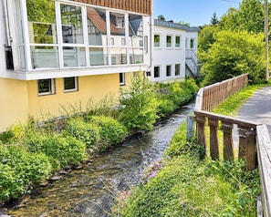 A narrow water channel with greenery on both sides, a wooden fence, and a building with multiple windows and a staircase.