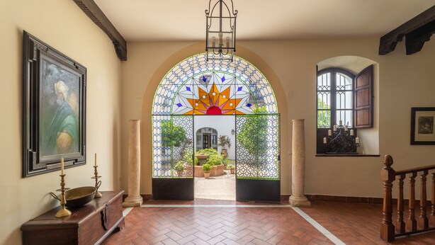 Un arco de entrada con un vitral, un banco de madera con un cuenco y candelabros, y un piso de baldosas.