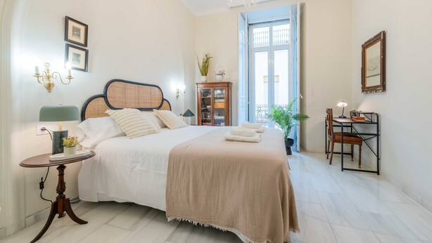A bedroom with a bed, bedside table, chandelier, and a view of the outside.