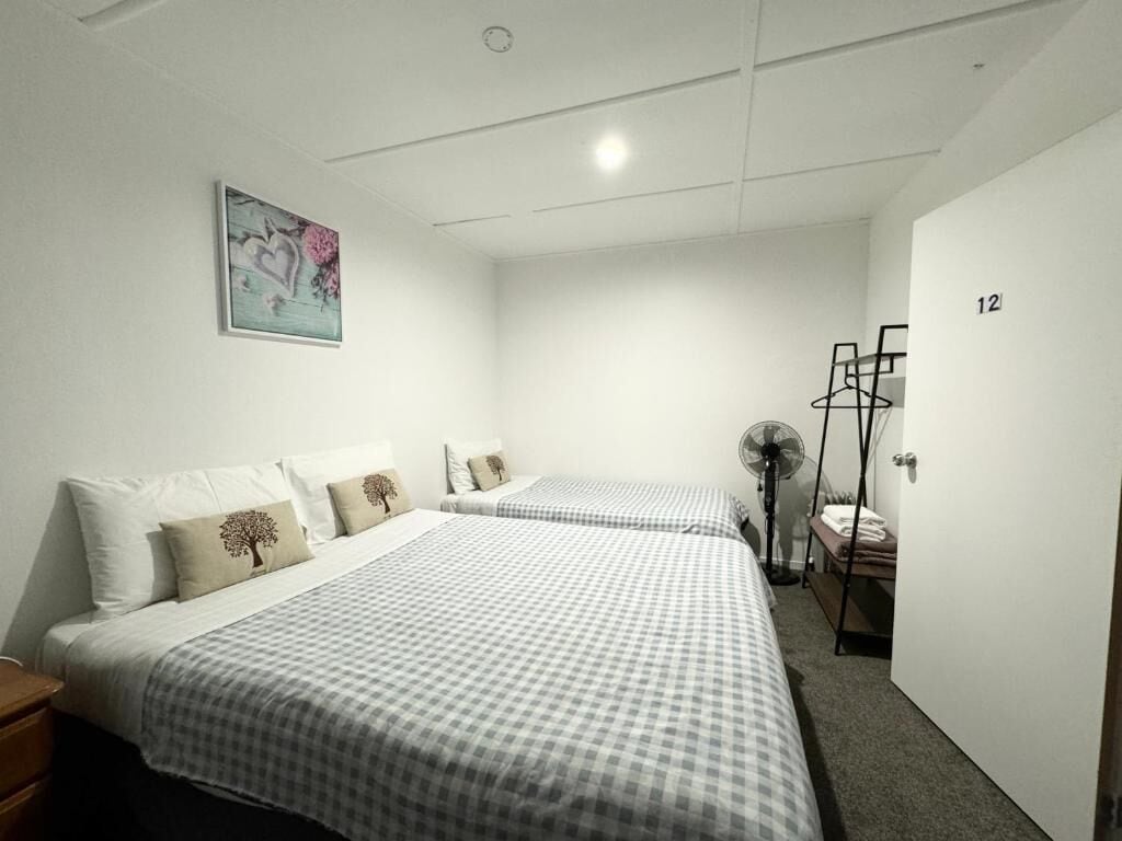 Quarto de hotel com duas camas, uma mesinha de cabeceira, uma obra de arte na parede, um cabideiro e uma porta com número.